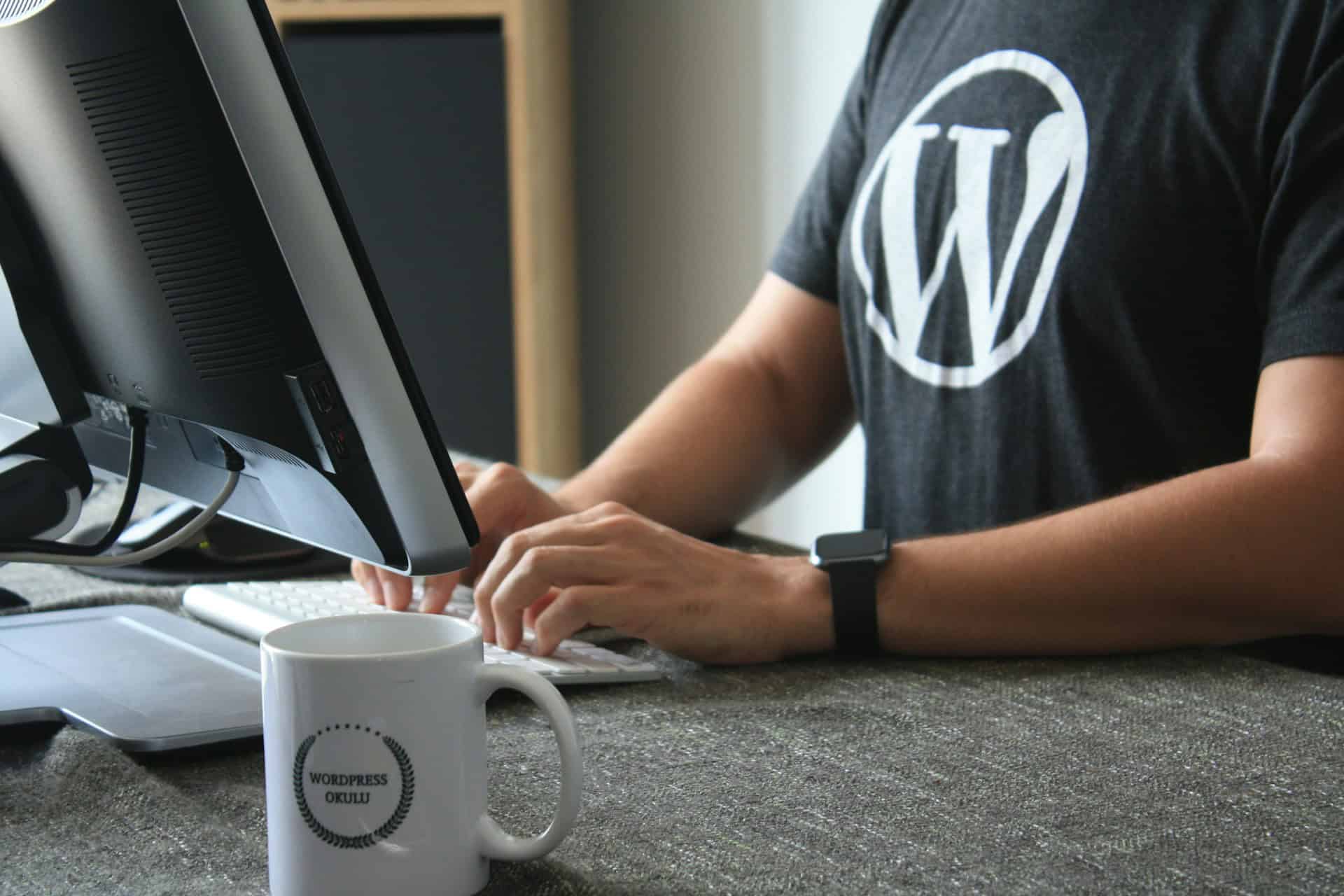 A person is typing at a desk with a desktop computer, wearing a T-shirt featuring the WordPress logo. On the table is a mug with the WordPress logo and "WORDPRESS GURU." The person, possibly engaged in professional WordPress maintenance, is wearing a smartwatch. The desk is covered with a dark cloth and the background is blurred.